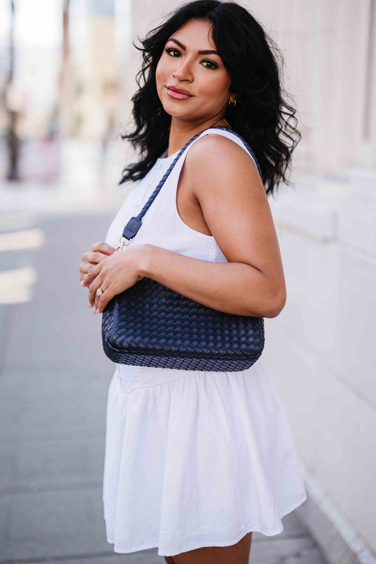 Woman in white dress holding a blue A woman wearing Drury Road Elle Woven Crossbody Bag for Women over her shoulder outdoors on a street.
