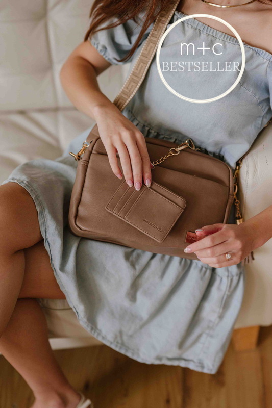 Woman holding a fawn colored Drury Road Alice Crossbody Vegan Leather bag, with the matching wallet and included key leash in hand with 'm+c BESTSELLER' label at the top right of the image.