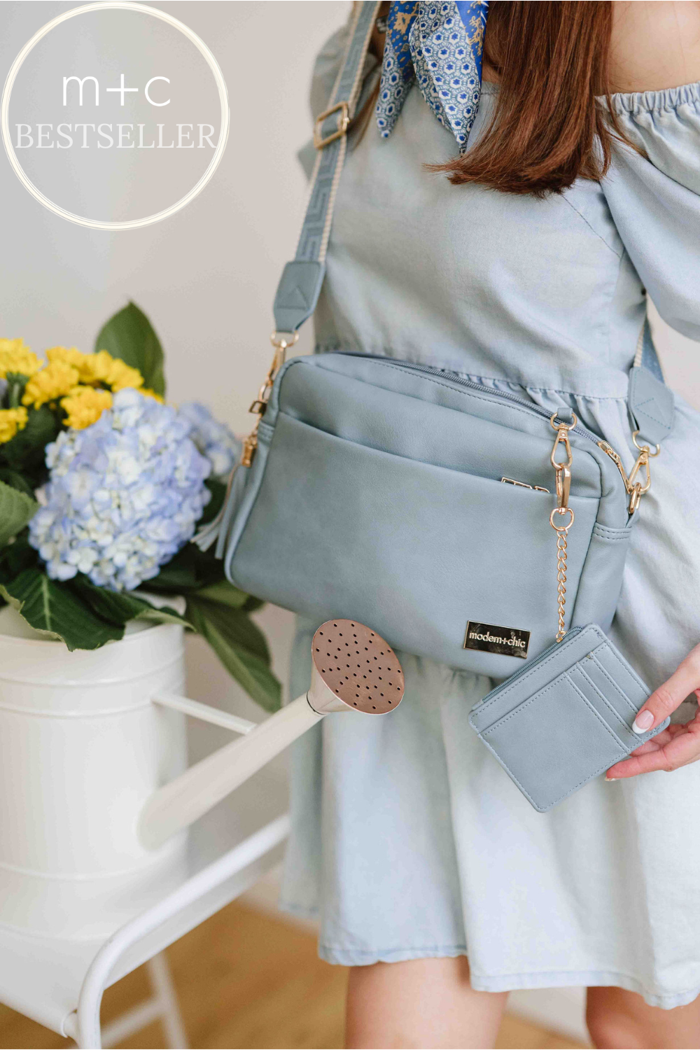 Woman wearing a blue dress wearing a dusty blue Drury Road Alice Crossbody Vegan Leather bag.