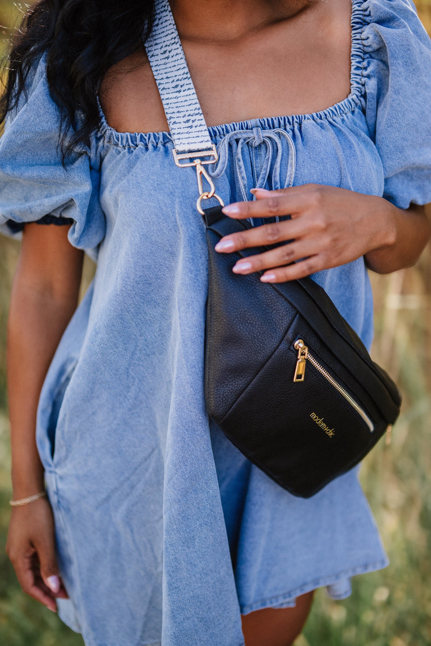 A woman wearing an off the should denim dress with a black Drury Road vegan leather Presley sling bag for women with coordinating blue printed strap crossbody. 