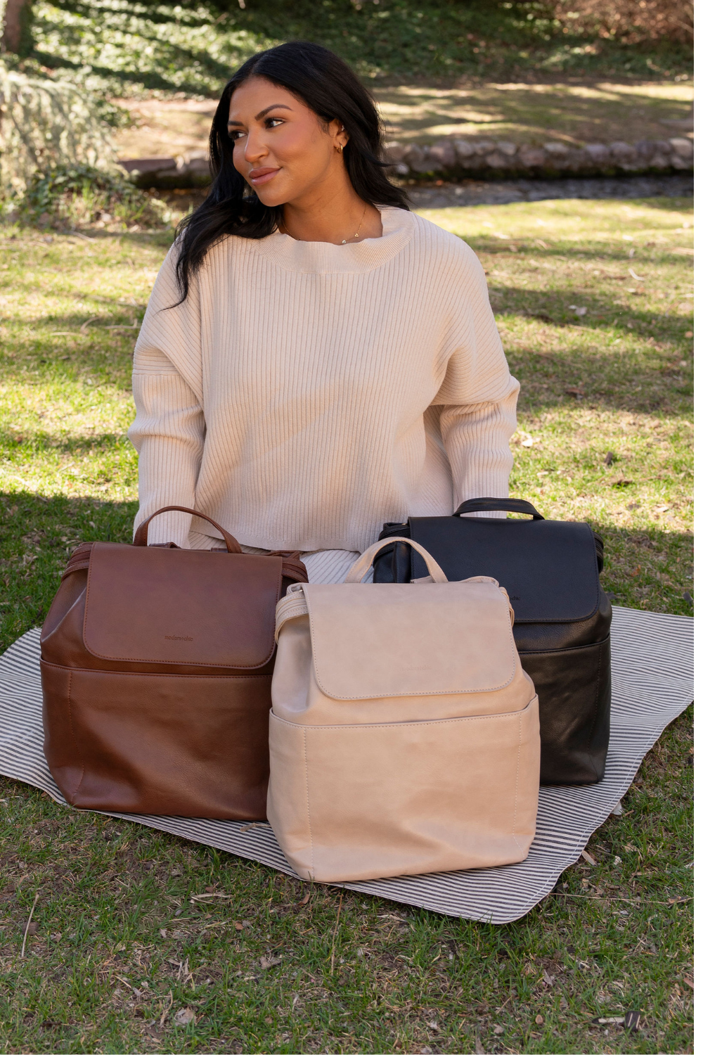 A woman kneeling on a black and white striped blanket sitting on the grass with three different Drury Road Brielle 2.0 Vegan Leather Convertible Backpack Purses for Women sitting in front of her in the  shades dark brown, taupe and black.