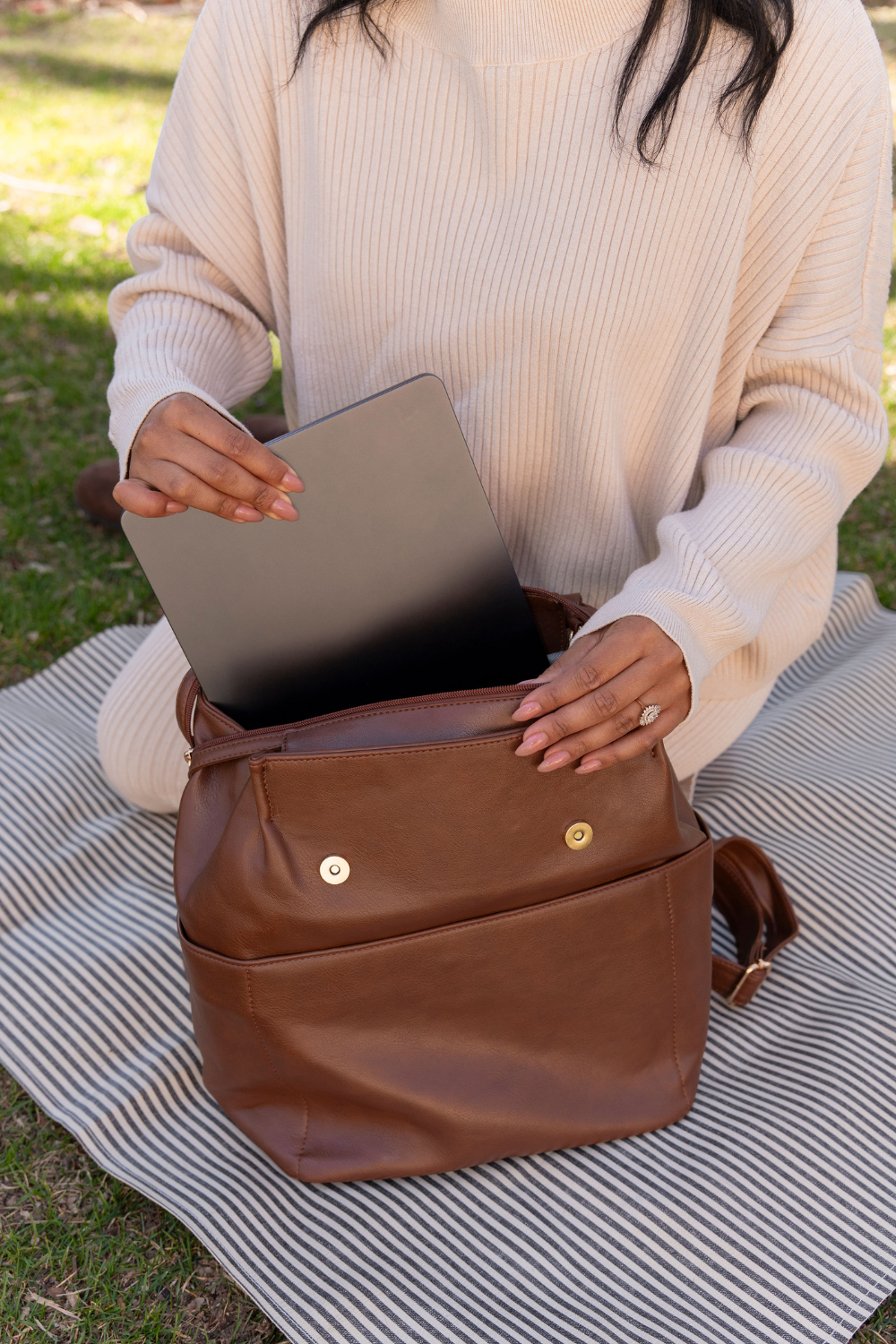 A woman kneeling on a black and white striped putting a laptop into a dark brown Drury Road Brielle 2.0 Vegan Leather Convertible Backpack Purse for Women with gold hardware. 
