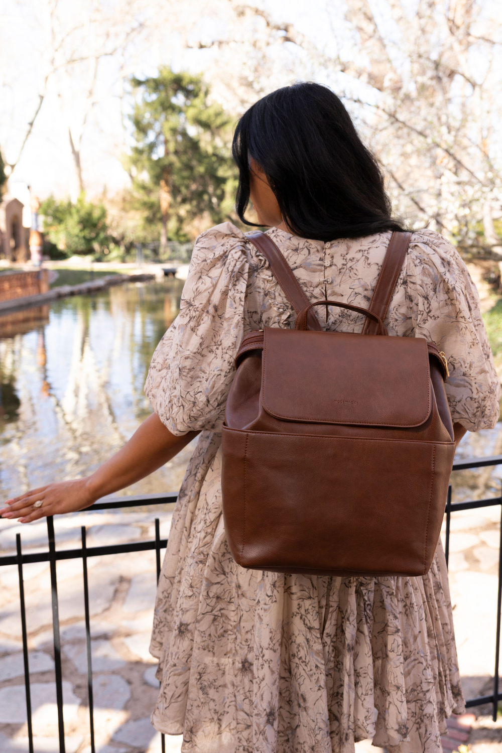 An image of a dark-haired woman facing away wearing a cream and brown floral dress with a dark brown Drury Road Brielle 2.0 Vegan Leather Convertible Backpack Purse for Women. 