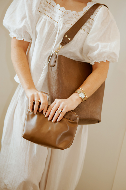 A woman holding a brown Drury Road vegan leather Sophie Bucket Bag and coordinating zipper pouch for Women –  with a blurred background.