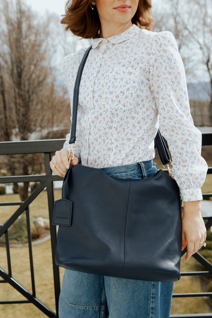 A woman wearing a floral white button up shirt with jeans and a black Drury Road Olivia Convertible Slouchy Tote Bag for Women across her body and over her hip.