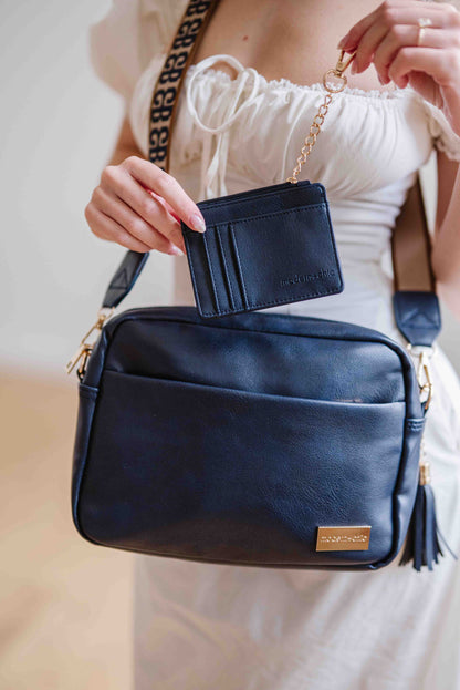 Women posing with a navy Drury Road Alice Crossbody vegan leather bag with gold hardware, matching key leash and wallet with a removable tassel attached.