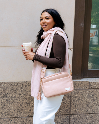 Woman holding a coffee cup, wearing a pink scarf and carrying a dusty blush Drury Road Alice Crossbody Vegan Leather bag  against a building background.