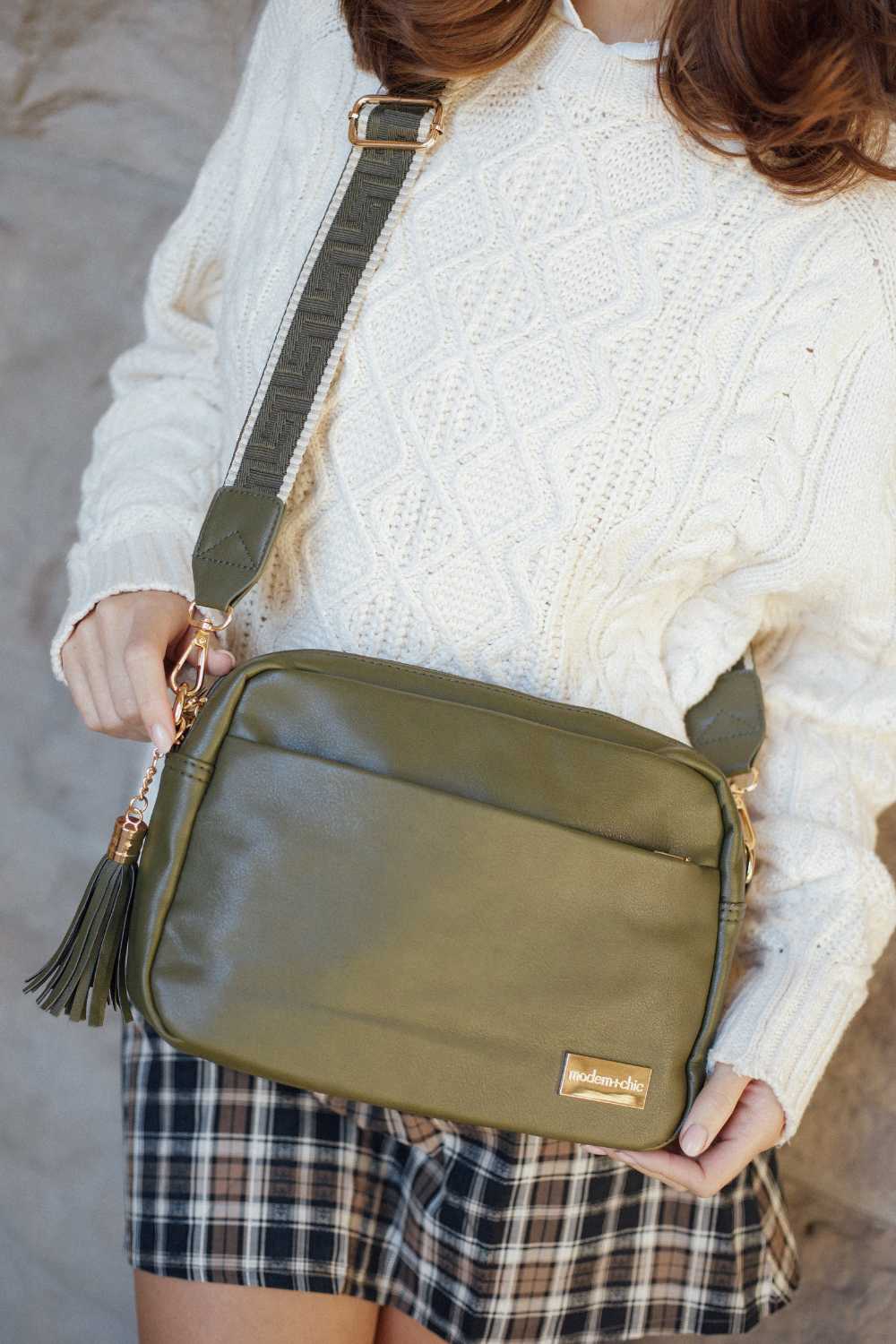 Person holding an olive green Drury Road Crossbody Vegan Leather Bag wearing a white sweater and plaid skirt.