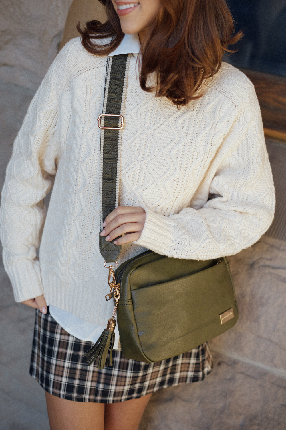 Person wearing a cream sweater and plaid skirt modeling an olive green Alice Crossbody Vegan leather bag. 
