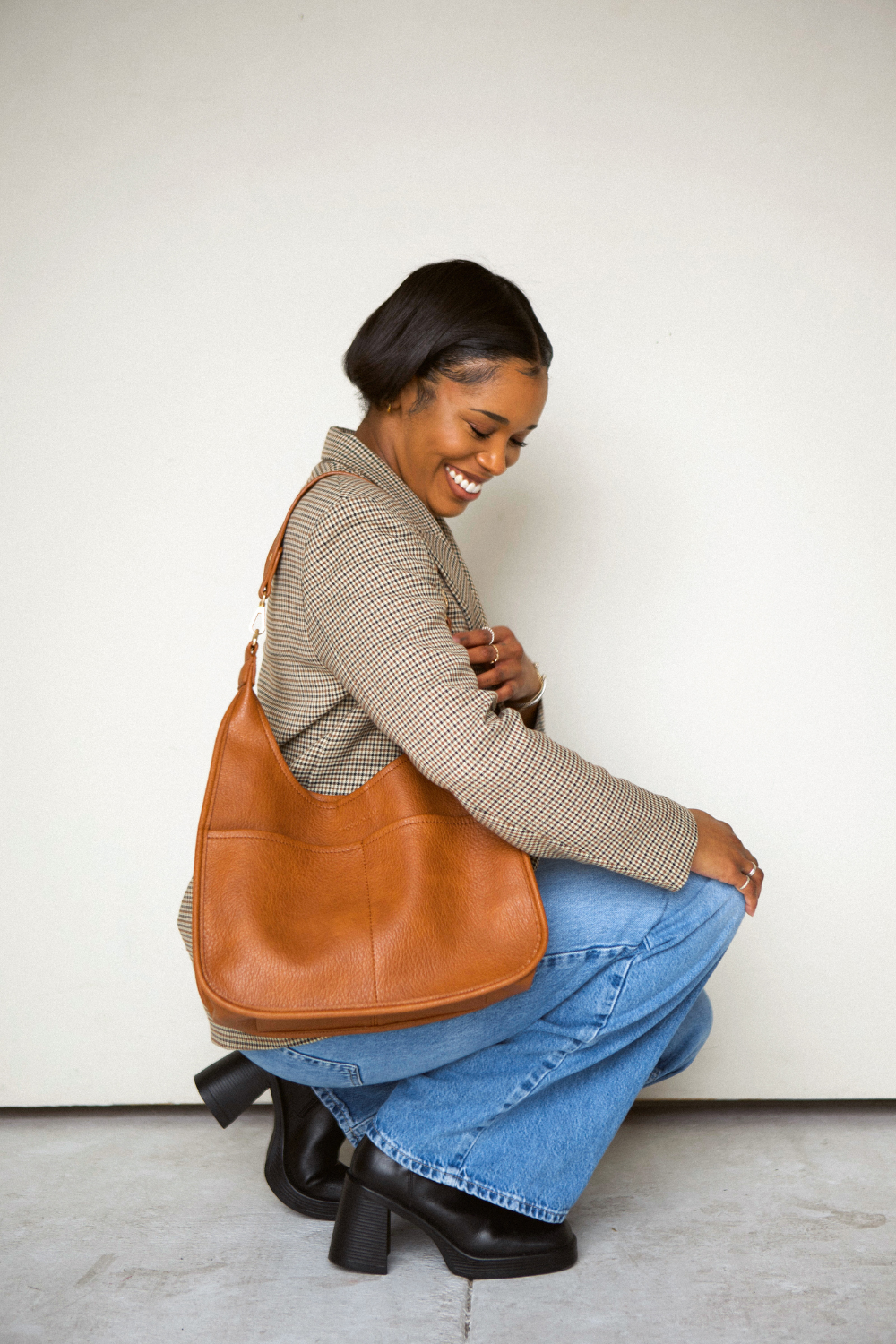 Woman squatting wearing a camel Drury Road Clara Vegan Leather Slouchy Tote Bag for Women against a white wall. 
