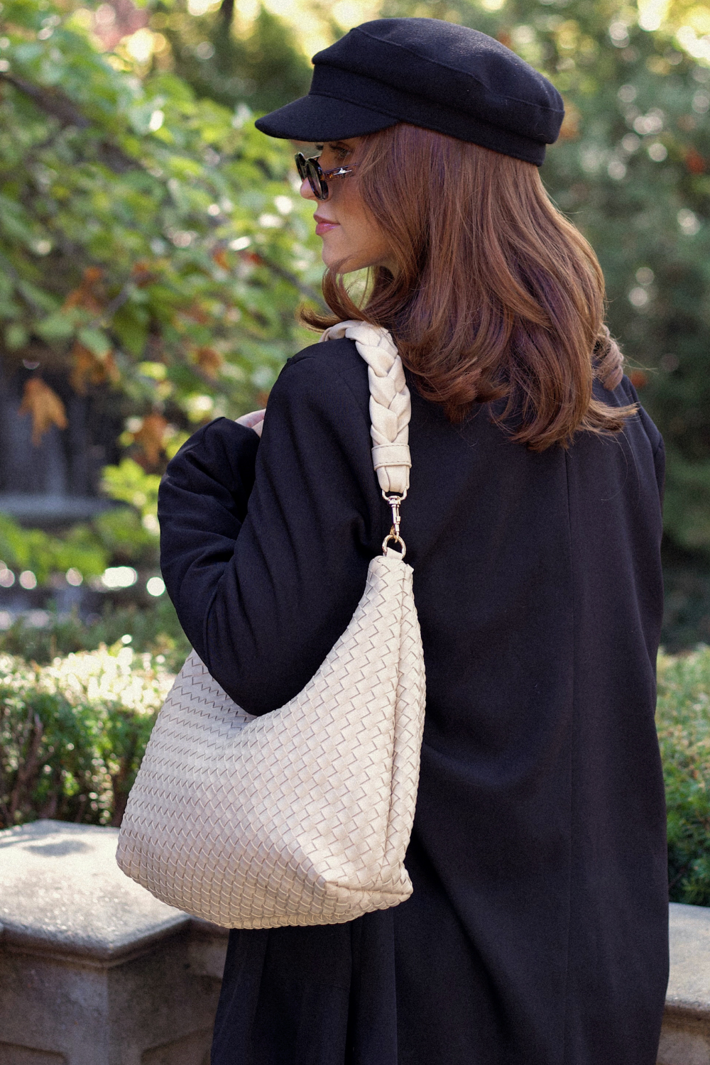 Woman in black coat and cap holding an ivory Drury Road Kelly Woven Convertible Crossbody Shoulder Bag outdoors.