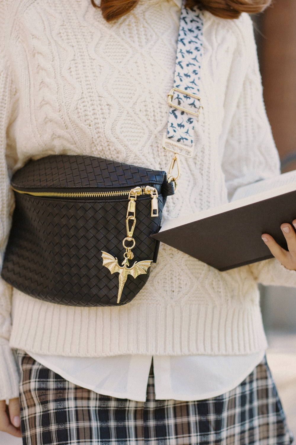 Person wearing a black textured bag with a gold dragon charm, a black and white dragon bag strap, holding a book, against a neutral background.