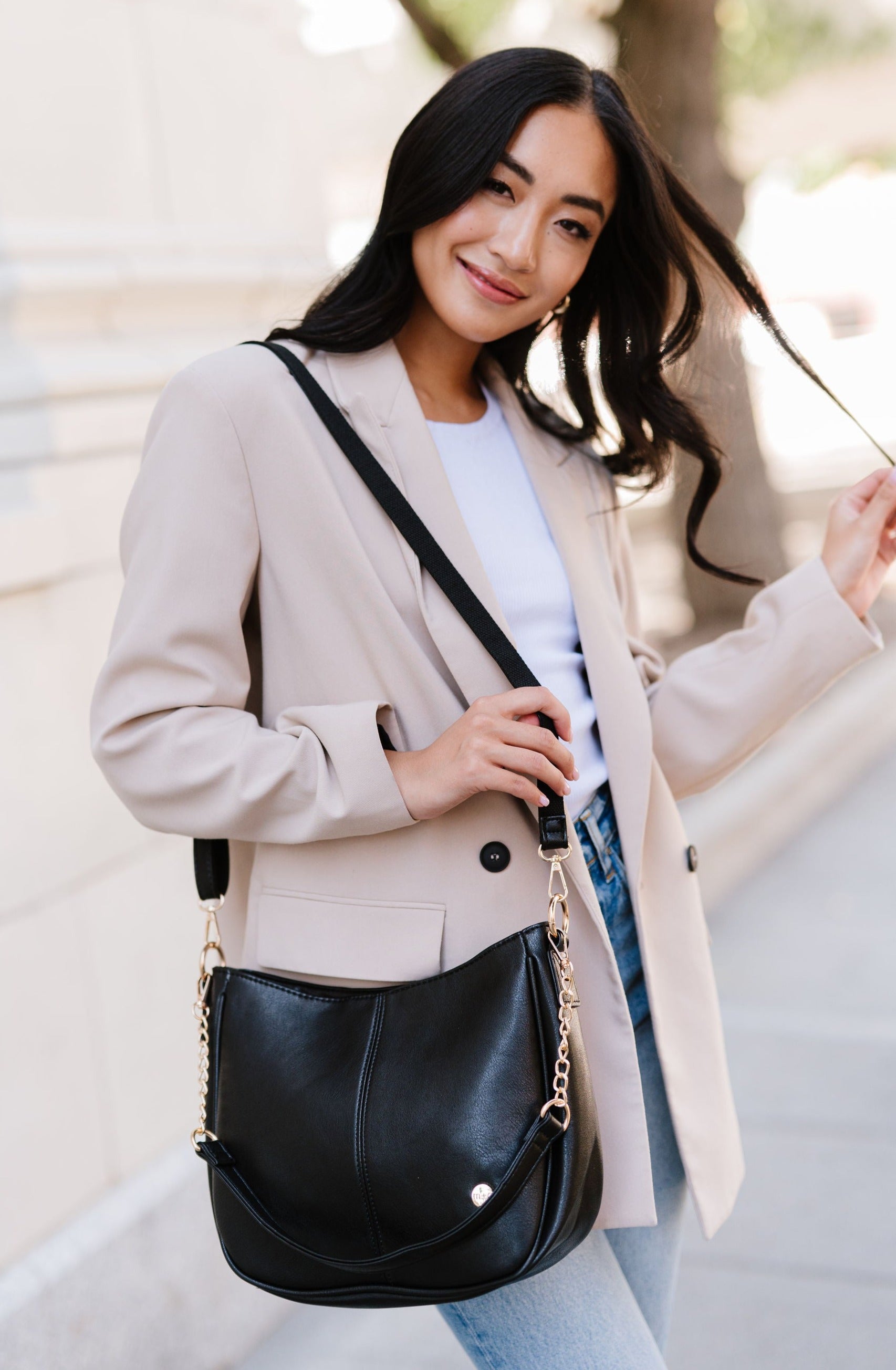 A woman standing in front of a white building wearing a black Drury Road Marissa vegan leather crossbody handbag.