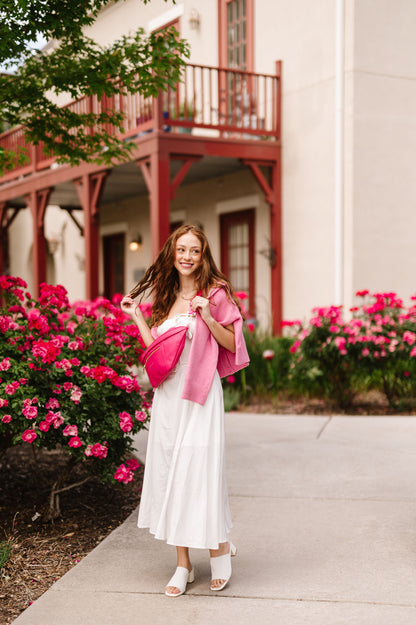 A woman wearing a white dress and pink sweater draped over her shoulders with a hot pink oversized Drury Road Presley Oversized Sling Bag for Women with gold hardware across her torso.