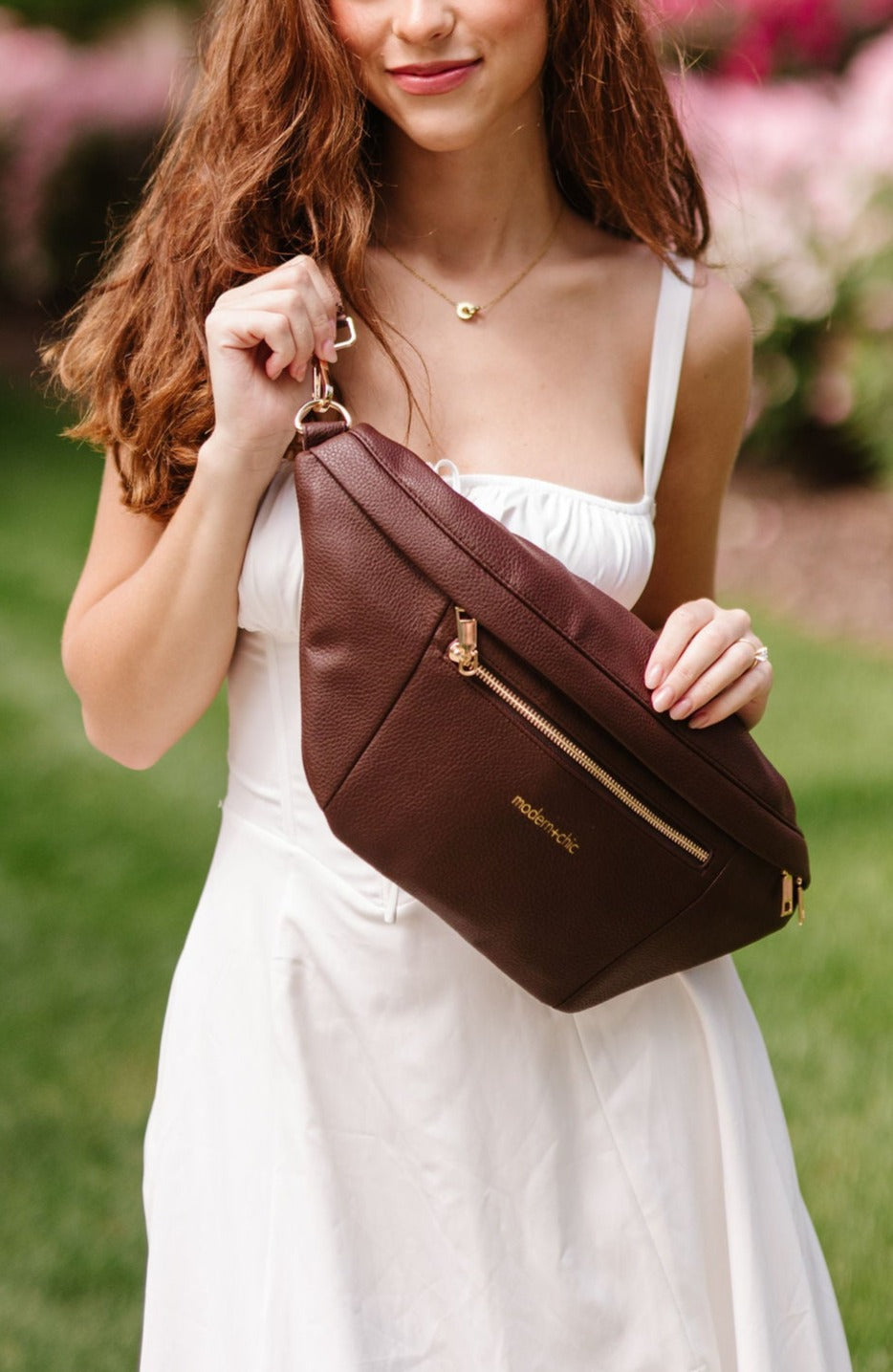 A woman wearing a white dress holding an oversized brown Drury Road Presley Oversized Sling Bag for Women with gold hardware as she wears it crossbody.