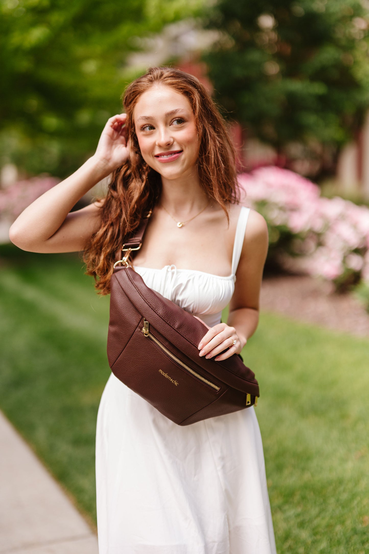 A woman wearing a white dress holding an oversized brown Drury Road Presley Oversized Sling Bag for Women with gold hardware as she wears it crossbody.