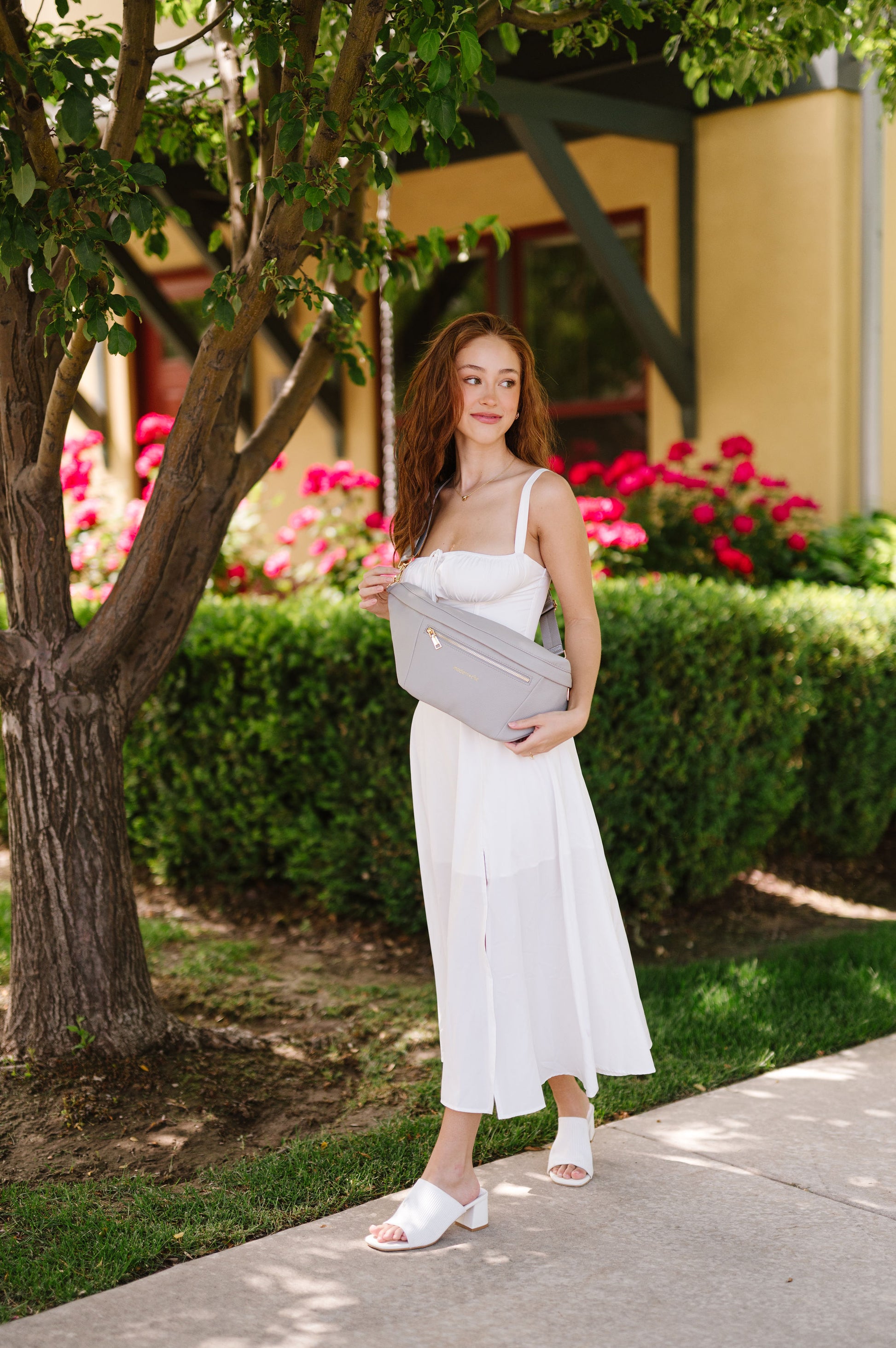 A woman wearing a white dress and an oversized grey Drury Road Presley Oversized Sling Bag for Women with gold hardware across her torso.
