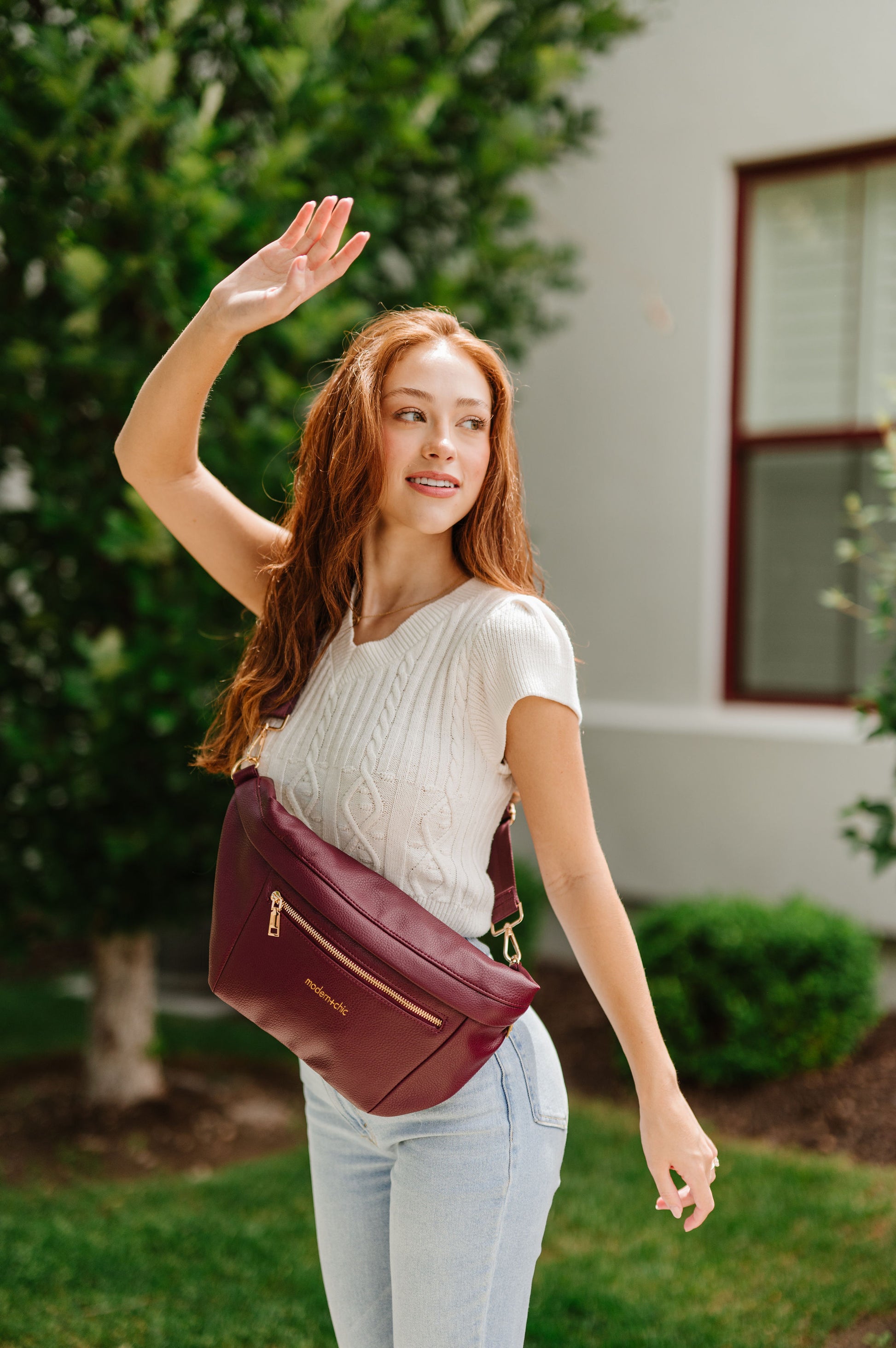 A woman wearing a white cropped sweater and an oversized wine colored Drury Road Presley Oversized Sling Bag for Women with gold hardware across her torso.