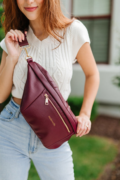 A woman wearing a white cropped sweater and an oversized wine colored Drury Road Presley Oversized Sling Bag for Women with gold hardware across her torso.