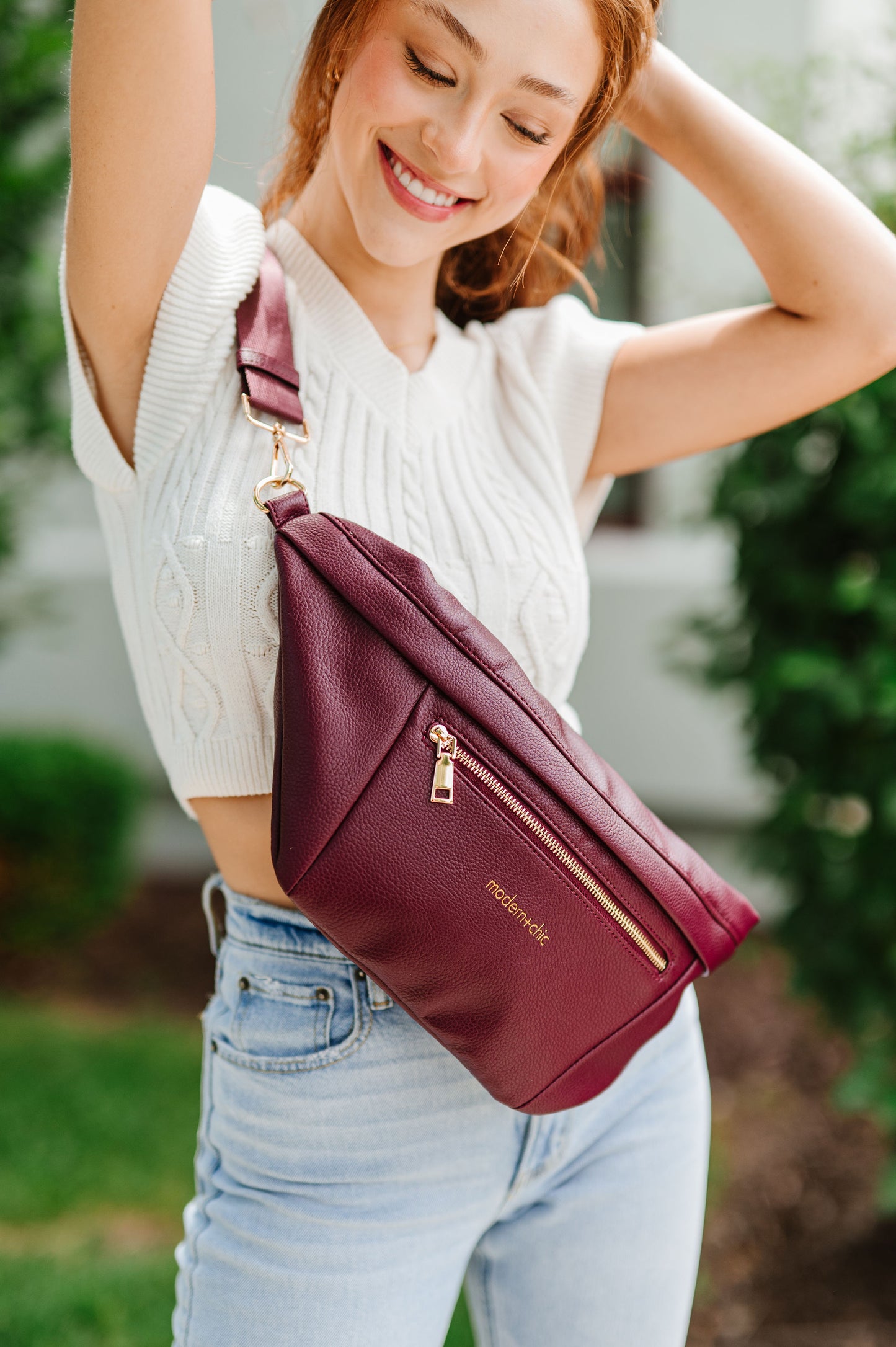 A woman wearing a white cropped sweater and an oversized wine colored Drury Road Presley Oversized Sling Bag for Women with gold hardware across her torso.