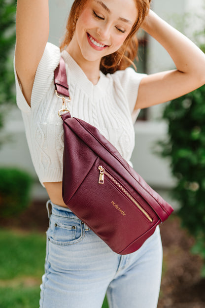 A woman wearing a white cropped sweater and an oversized wine colored Drury Road Presley Oversized Sling Bag for Women with gold hardware across her torso.