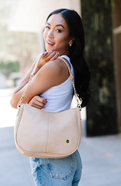 A woman wearing a cream Drury Road Marissa Vegan Leather Crossbody handbag over her shoulder displaying the gold chain accents on the strap.