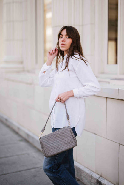 Woman in white shirt and jeans holding a gray Drury Road Elle Woven Shoulder Bag for Women against a light-colored building.