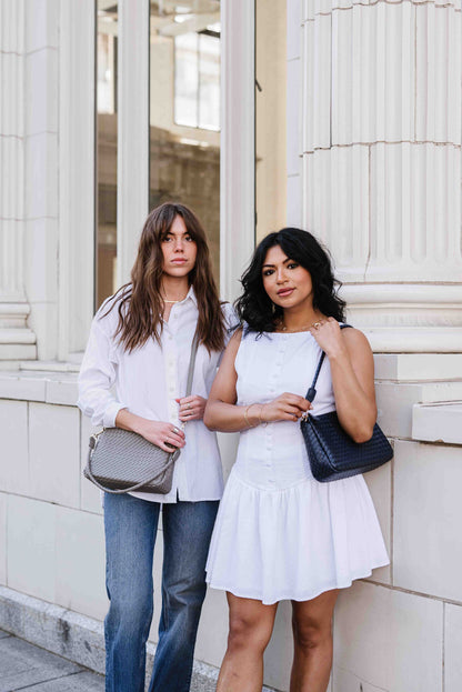 Two women standing next to each other, one holding a gray Drury Road Elle Woven Crossbody Bag for Women over crossbody and the other holding a black one. Both are wearing white and the setting appears to be outdoors with columns in the background.