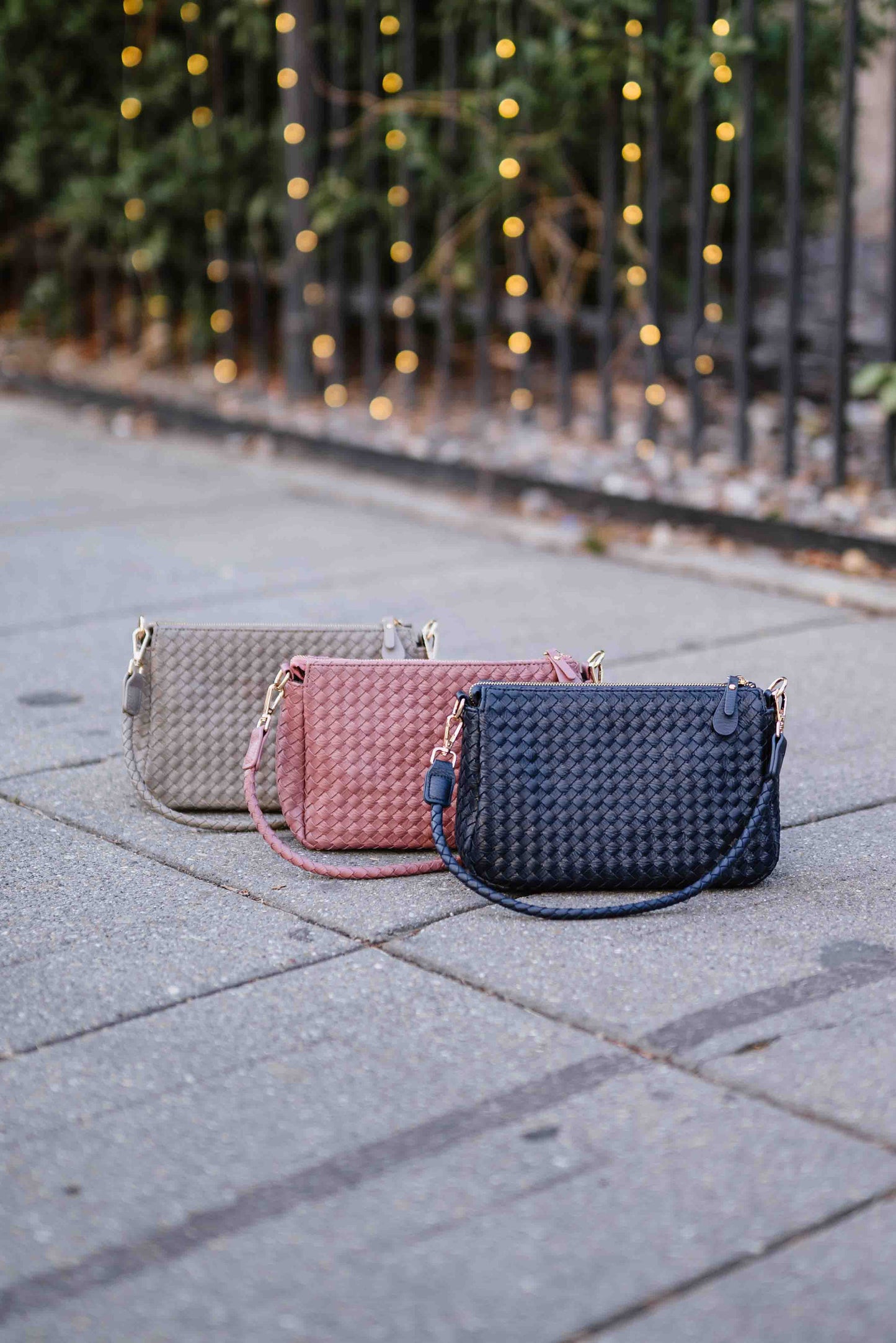 Three woven Drury Road Elle Woven Crossbody Bag  in gray, pink, and blue on a paved surface with blurred lights in the background.