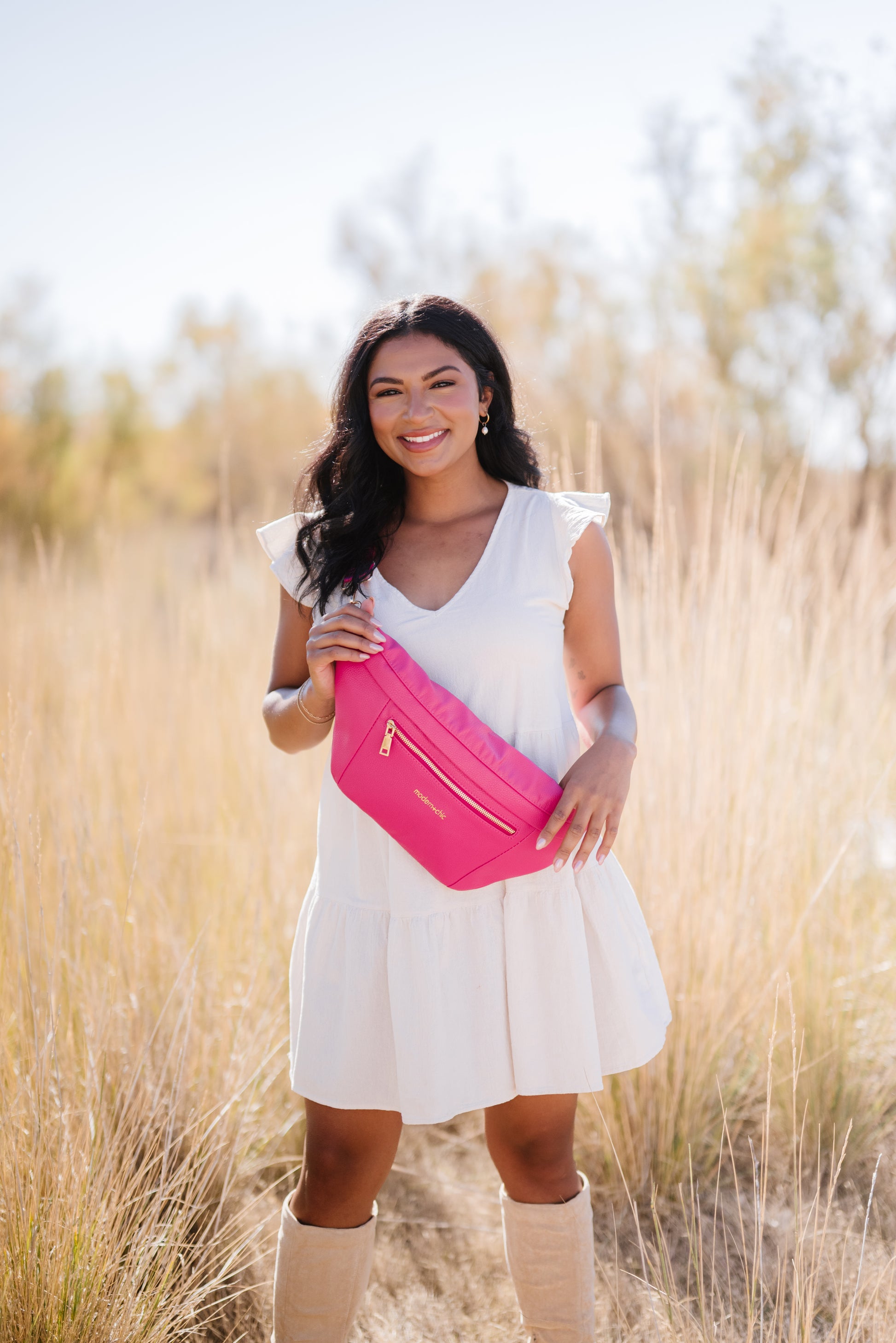 A woman wearing a white dress and tall boots with a hot pink oversized Drury Road Presley Oversized Sling Bag for Women with gold hardware across her torso.
