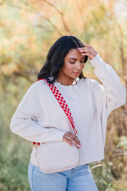 A woman wearing a light pink sweater and blue jeans is shown with a red and pink Drury Road Lizzie checkered adjustable strap attached to a light pink purse.