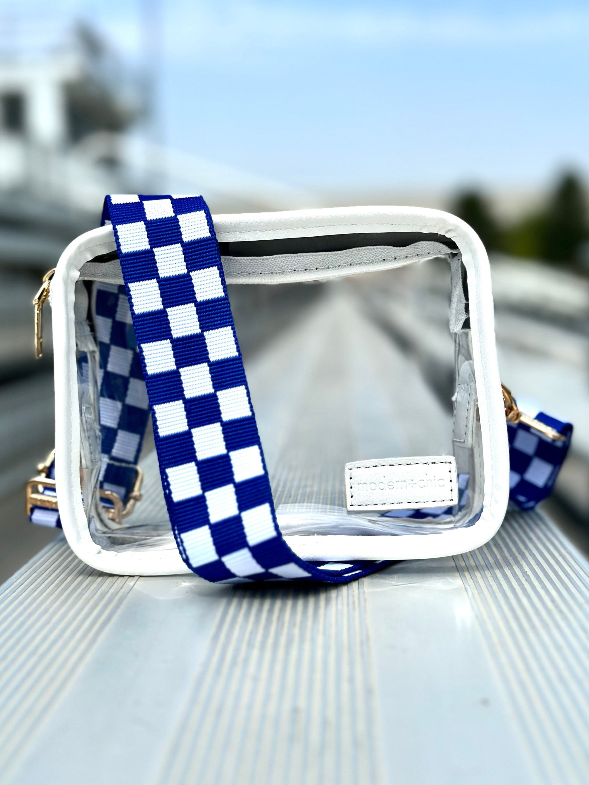 A clear handbag wrapped in a blue and white Drury Road Lizzie Checkered Purse Strap for Crossbody Bags sitting on an empty bleacher. 