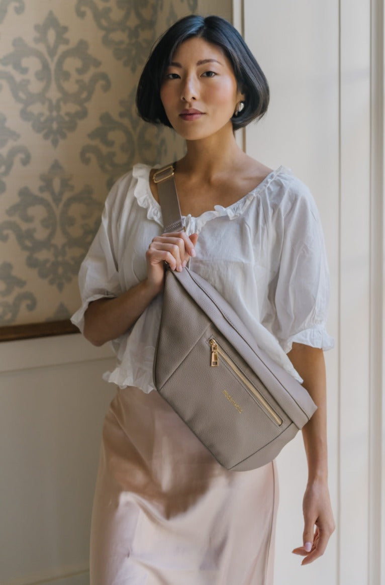 A woman wearing an oversized taupe Drury Road Presley Oversized Sling Bag for Women with gold hardware across her torso.