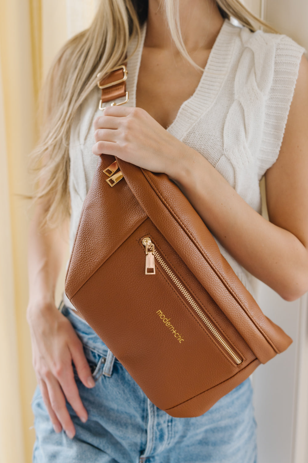 A woman wearing a white sweater vest and an oversized camel Drury Road Presley Oversized Sling Bag for Women with gold hardware across her torso.
