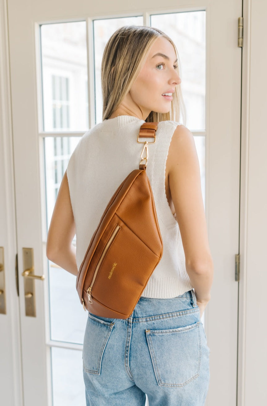 A woman wearing a white sweater vest and an oversized camel Drury Road Presley Oversized Sling Bag for Women with gold hardware across her back.