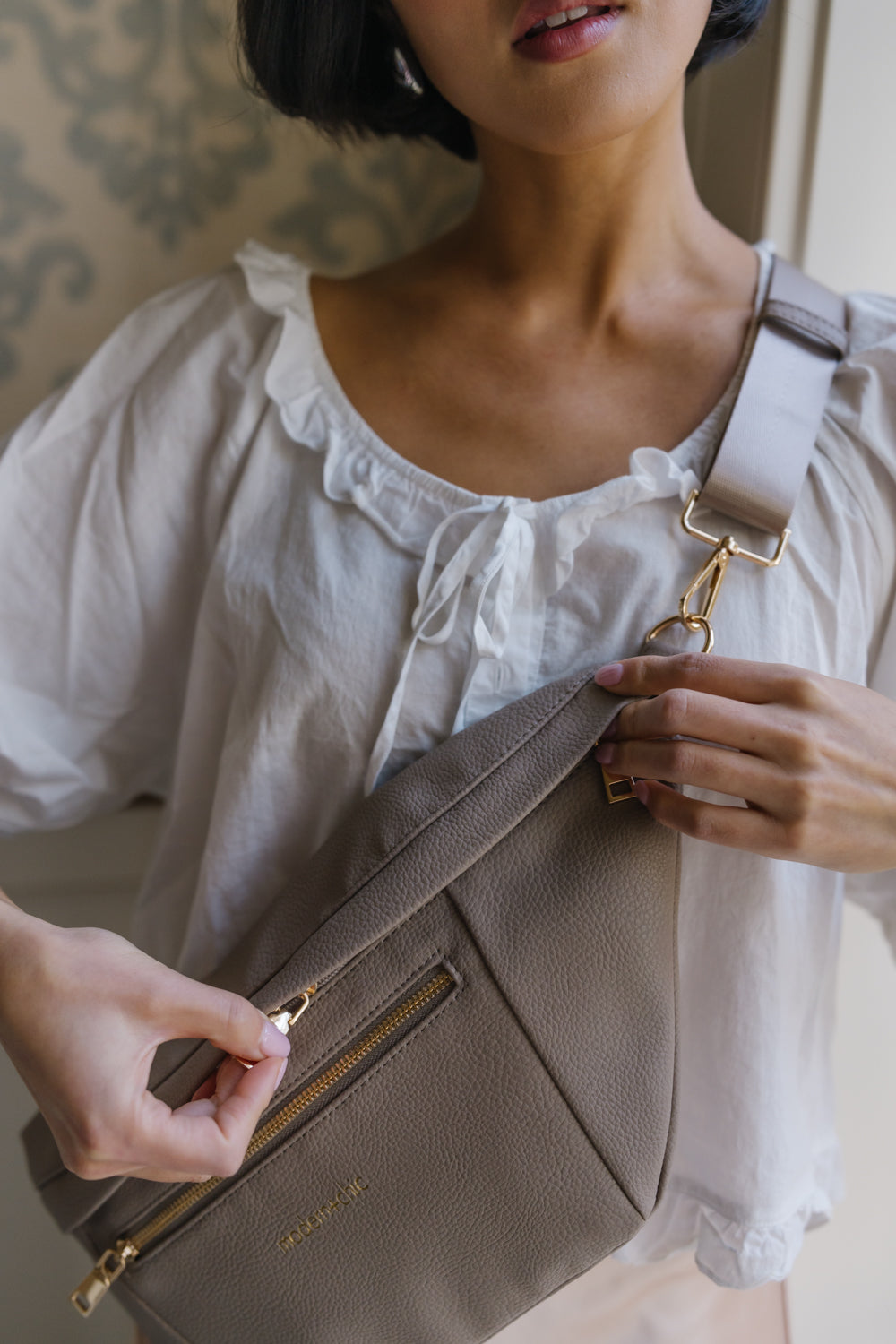A woman wearing an oversized taupe Drury Road Presley Oversized Sling Bag for Women with gold hardware across her torso.