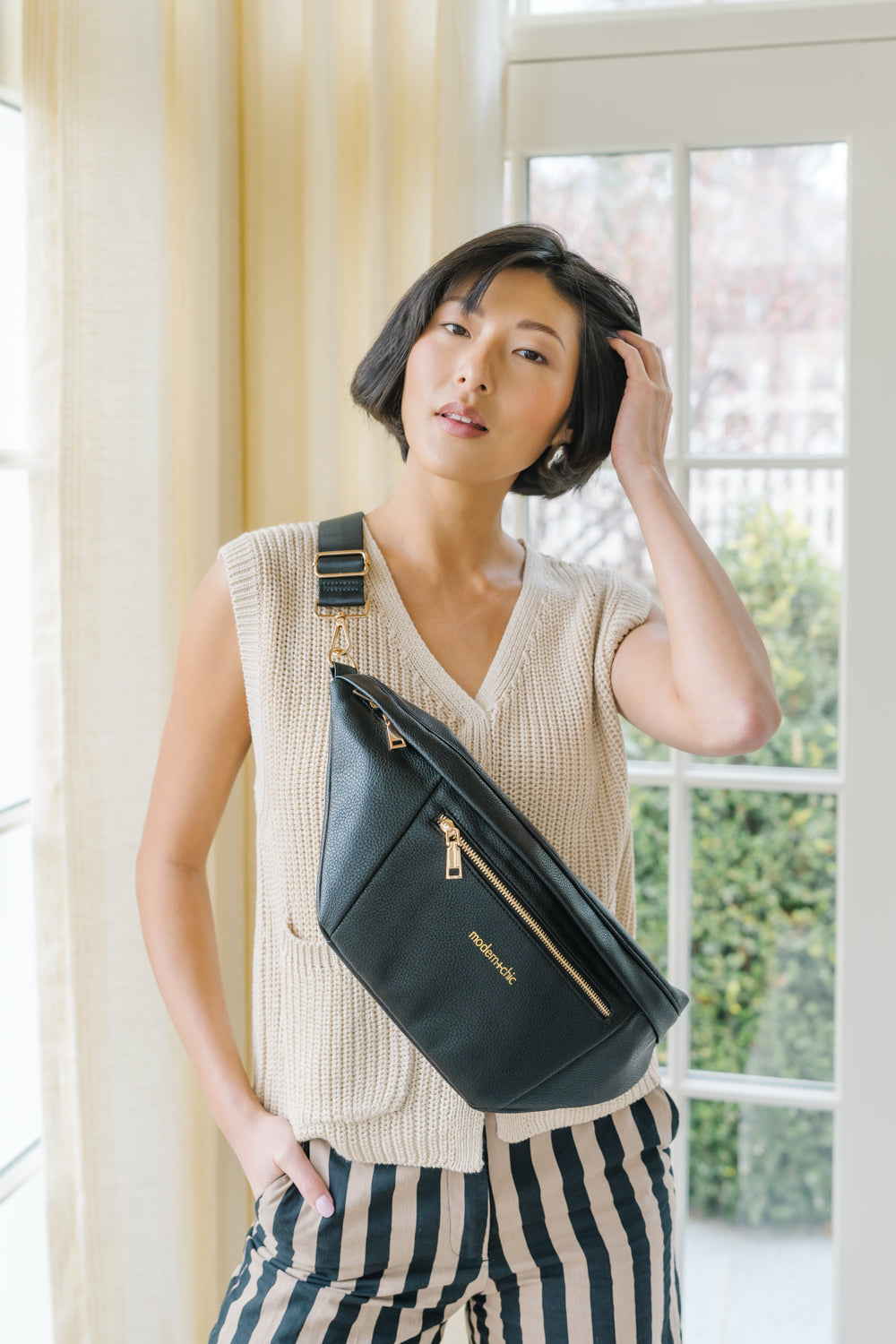 A woman wearing a cream sweater vest and an oversized black Drury Road Presley Oversized Sling Bag for Women with gold hardware across her torso.