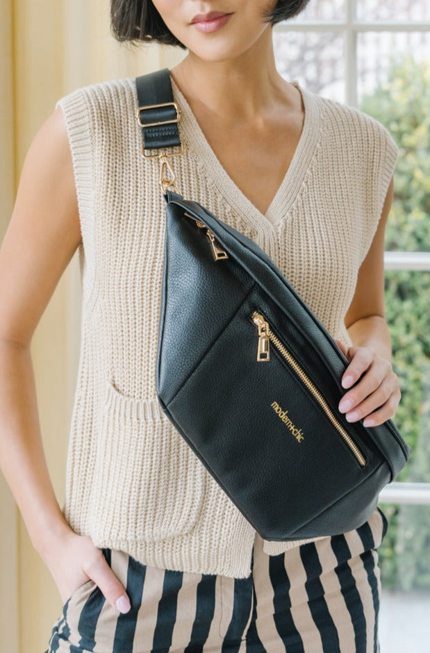 A woman wearing a cream sweater vest and an oversized black Drury Road Presley Oversized Sling Bag for Women with gold hardware across her torso.