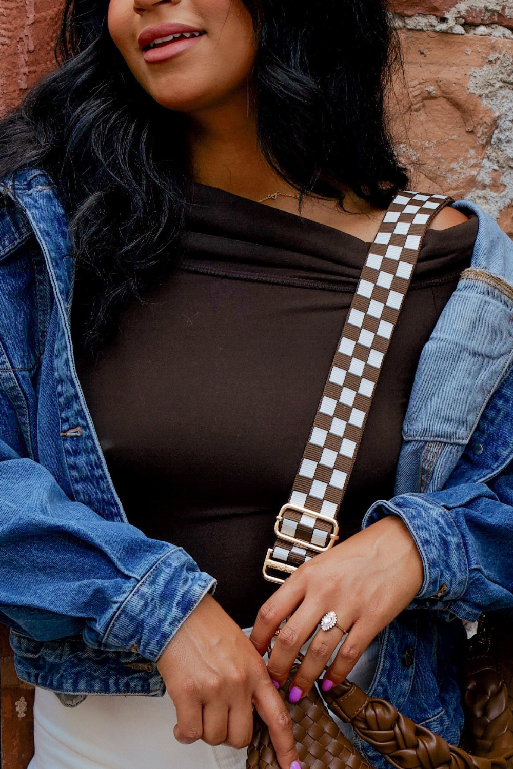 Woman wearing a denim jacket and brown top with a brown and white Drury Road Lizzie checkered adjustable strap bag against a brick wall.