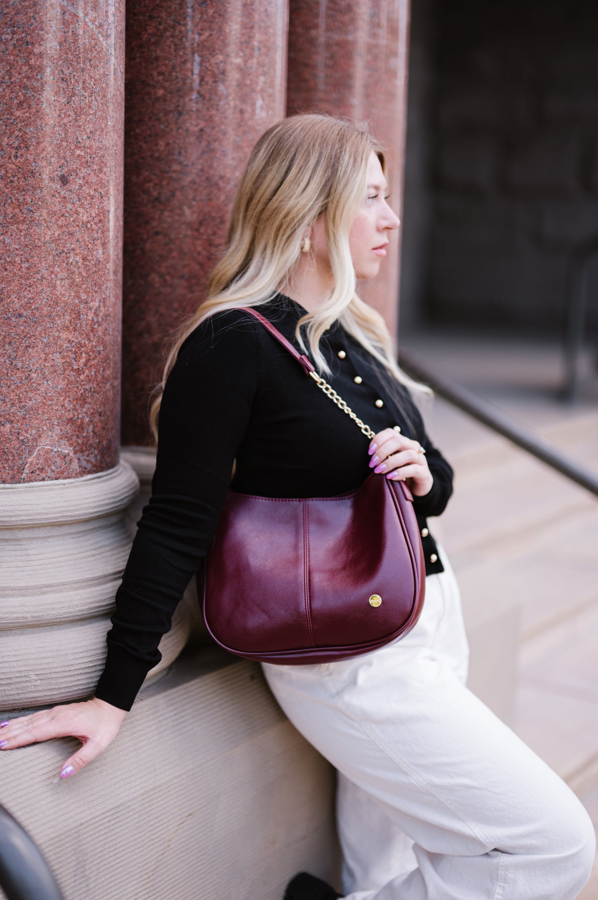 Woman leaning against a wall with a wine Drury Road Marissa vegan leather handbag on her should displaying gold chain accents on the strap. 