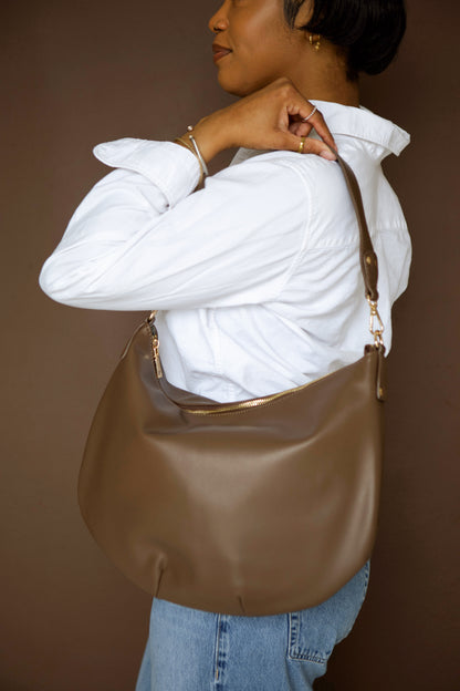 Person wearing a white shirt and blue jeans, holding a brown vegan leather Drury Road Savannah Slouchy Tote against a brown background. 