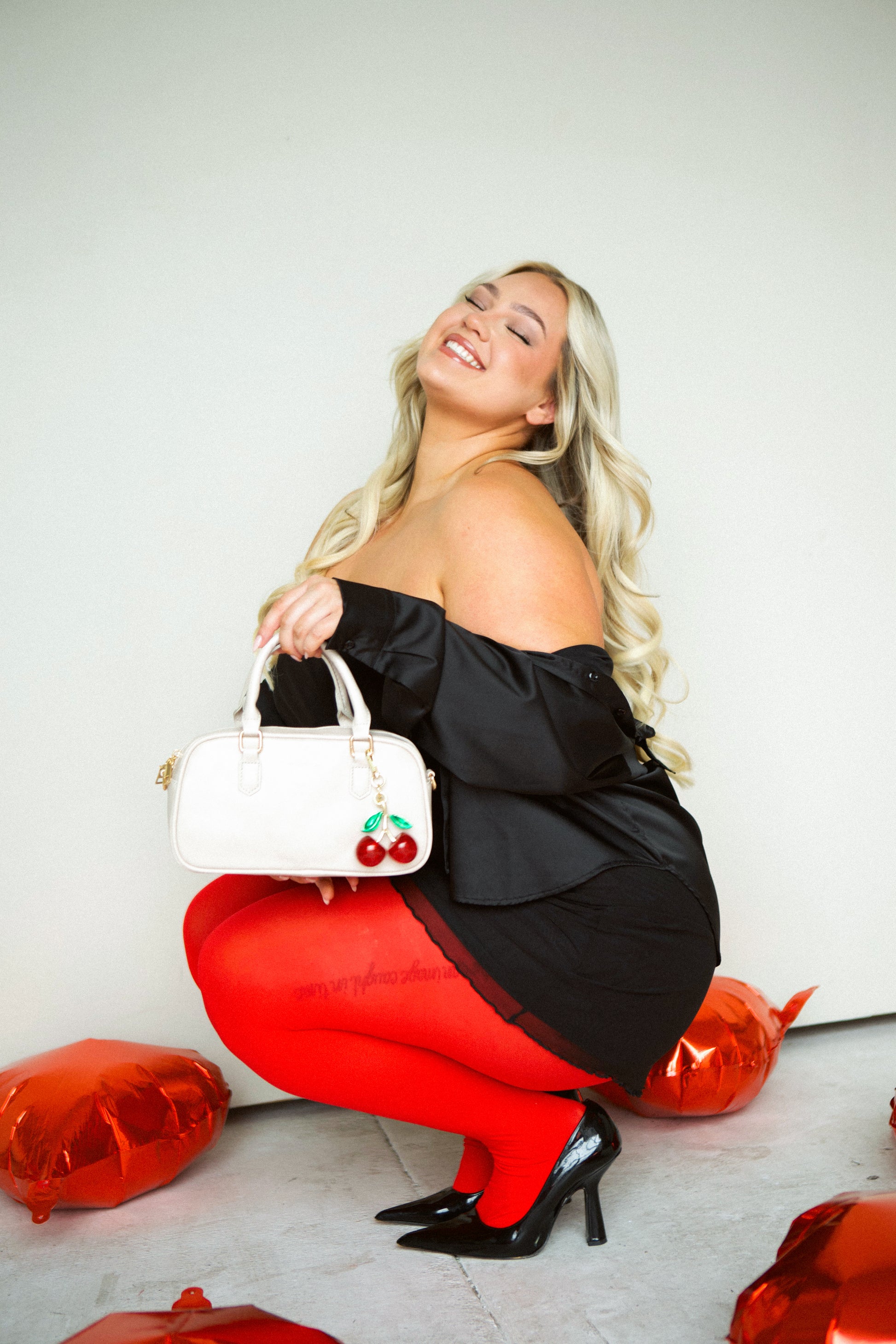  A woman wearing a black stress squatting down holding her cream Drury Road Remi Vegan Leather handbag with  and a large cherry charm hanging from the strap.
