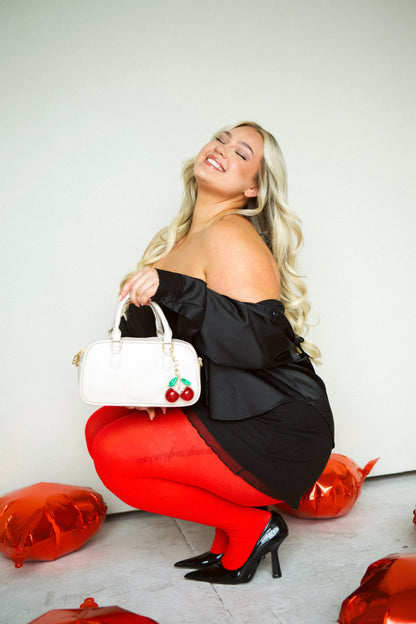  A woman wearing a black stress squatting down holding her cream Drury Road Remi Vegan Leather handbag with  and a large cherry charm hanging from the strap.
