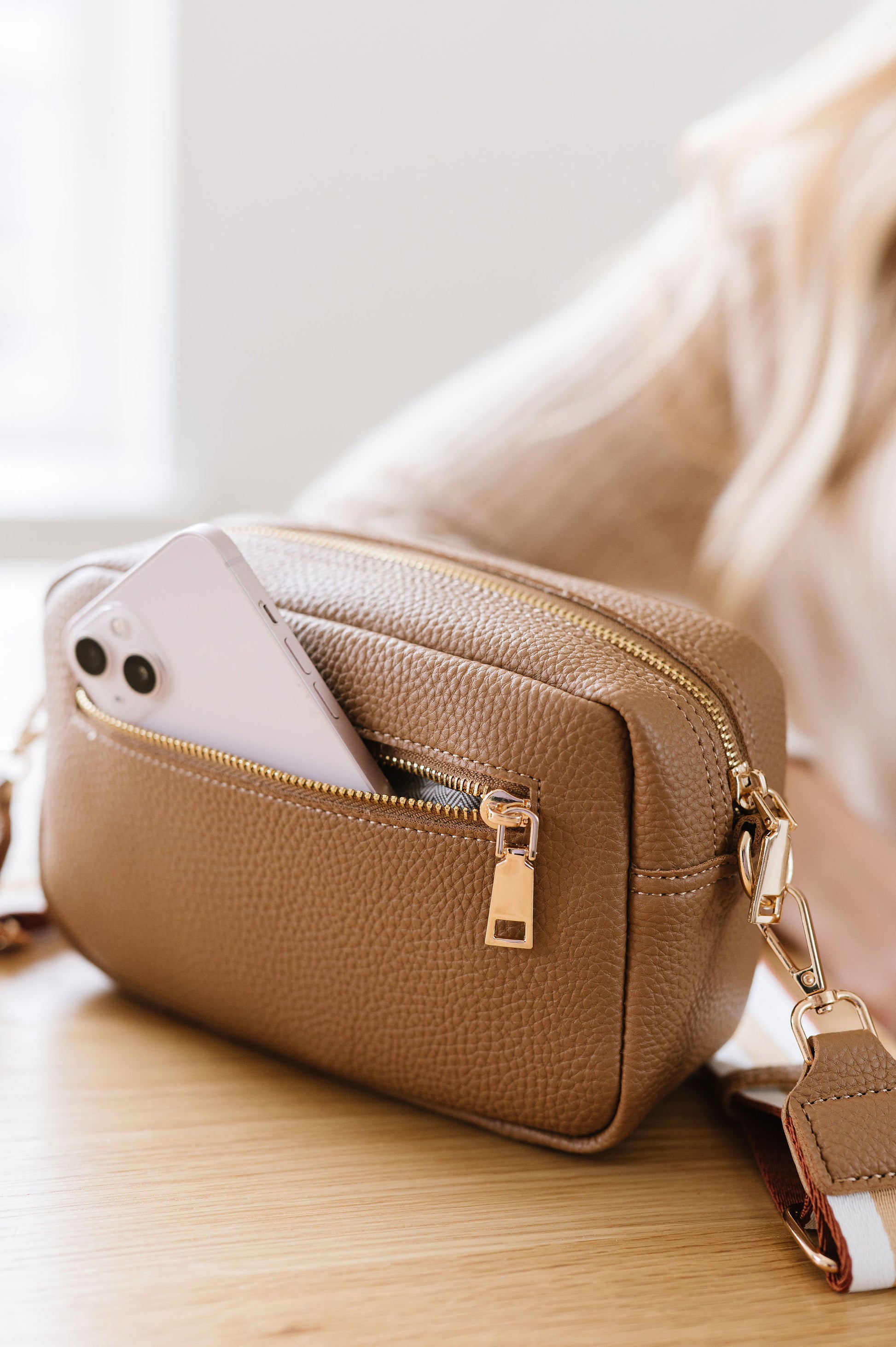 A beige Drury Road Willow Vegan Leather Convertible Crossbody Bag sitting on a table with a pink phone partially showing out of the unzipped back pocket. 