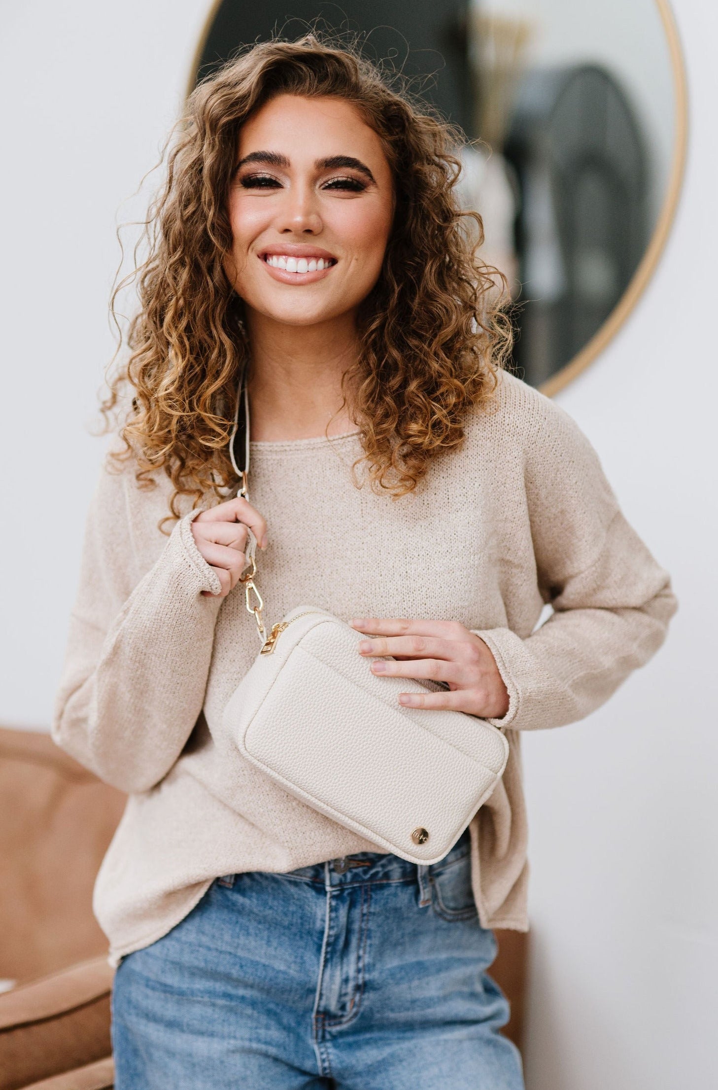 A woman with curly hair is smiling while wearing a beige sweater, blue jeans and a beige Willow Convertible Crossbody bag with gold hardware.