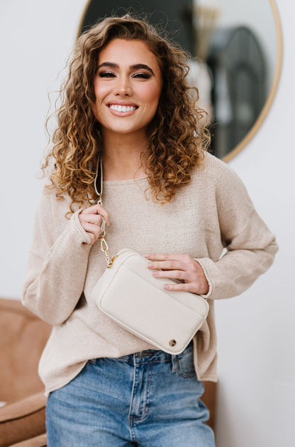 A woman with curly hair is smiling while wearing a beige sweater, blue jeans and a beige Willow Convertible Crossbody bag with gold hardware.